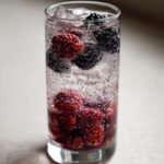 Close-up of a tall glass filled with ice, sparkling water, raspberries, and blackberries for a Blueberry Blackberry Mocktail.