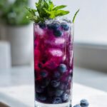 A tall glass filled with vibrant Blueberry Mint Lemonade, ice, fresh blueberries, and a mint sprig garnish.