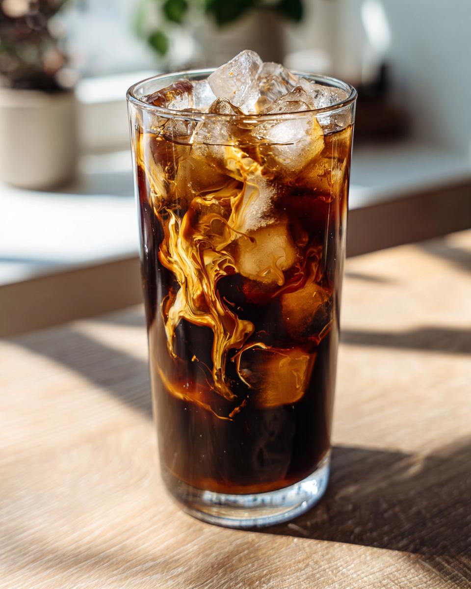 Close-up of a tall glass filled with ice cubes, dark coffee, and caramel swirling into the liquid, making a Caramel Iced Coffee.