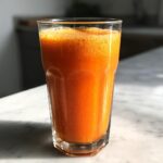 A tall glass filled with vibrant orange Carrot Ginger Citrus Turmeric Juice, showing foam on top, set on a marble surface.