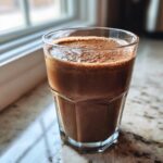 A close-up of a frothy Chocolate Hazelnut Smoothie served in a clear, faceted glass on a granite countertop.