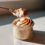 A spoonful of creamy topping being lifted from a jar of Cinnamon Roll Overnight Oats dusted with cinnamon.