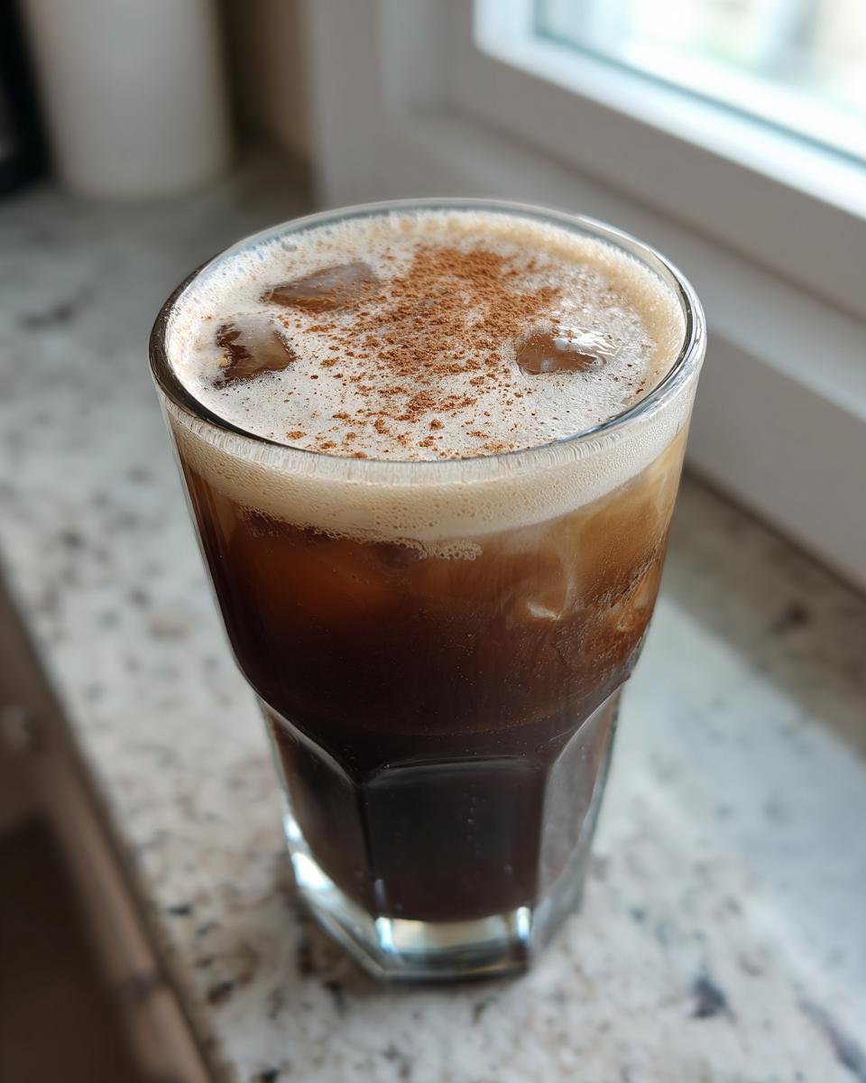 Close-up of a tall glass of Cinnamon Vanilla Iced Coffee topped with foam and sprinkled cinnamon.