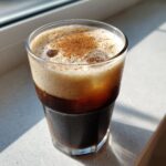 A tall glass of Cinnamon Vanilla Iced Coffee with ice, topped with creamy foam and a sprinkle of cinnamon.