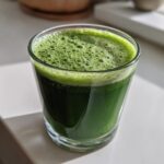Close-up of a rich green Cleansing Sugar Detox Juice in a clear glass, topped with fine foam.