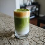 A tall glass of layered Coconut Almond Matcha Latte showing white milk, green matcha, and a foamy orange/yellow top dusted with matcha powder.