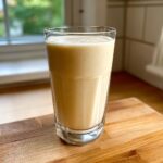 A tall glass filled with a thick, creamy Coconut Almond Smoothie resting on a wooden cutting board.