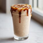 A tall glass filled with a thick Coconut Caramel Smoothie, topped with a rich caramel drizzle dripping down the sides.