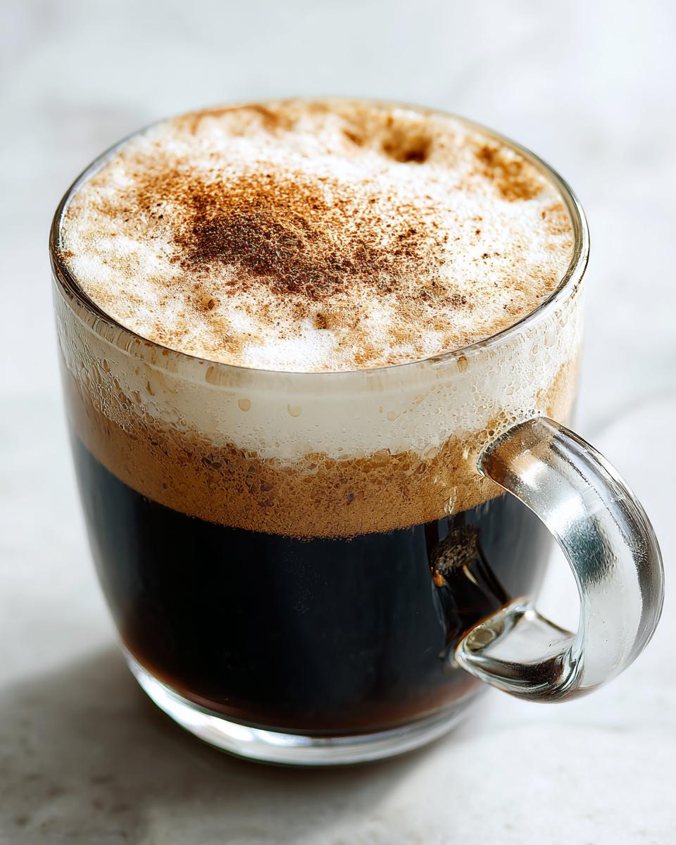 Close-up of a rich Coconut Chai Tea Latte with thick foam topped with cinnamon in a clear glass mug.
