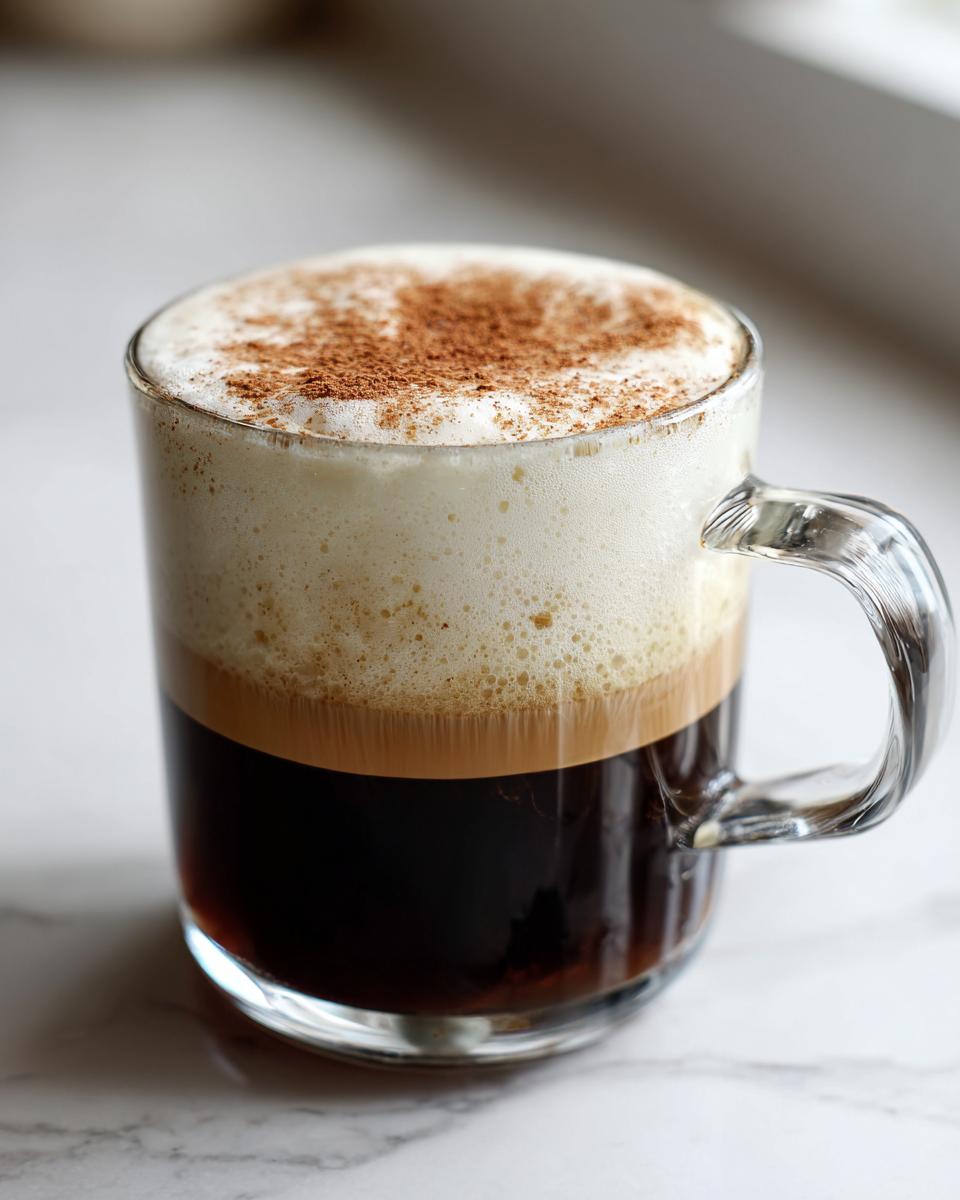 A clear glass mug filled with a layered Coconut Chai Tea Latte, topped with thick foam and sprinkled cinnamon