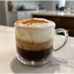 A layered Coconut Chai Tea Latte with thick foam topping dusted with cinnamon in a clear glass mug.