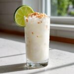 A tall, frosty glass of a creamy Coconut Lime Mocktail garnished with a lime wheel and toasted coconut flakes.