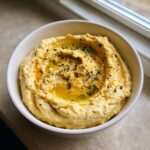 A close-up of creamy Roasted Garlic Hummus, drizzled with olive oil and sprinkled with herbs and paprika.