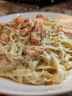 A close-up of a white plate piled high with Creamy Salmon Alfredo Pasta, topped with chunks of salmon and dried herbs.
