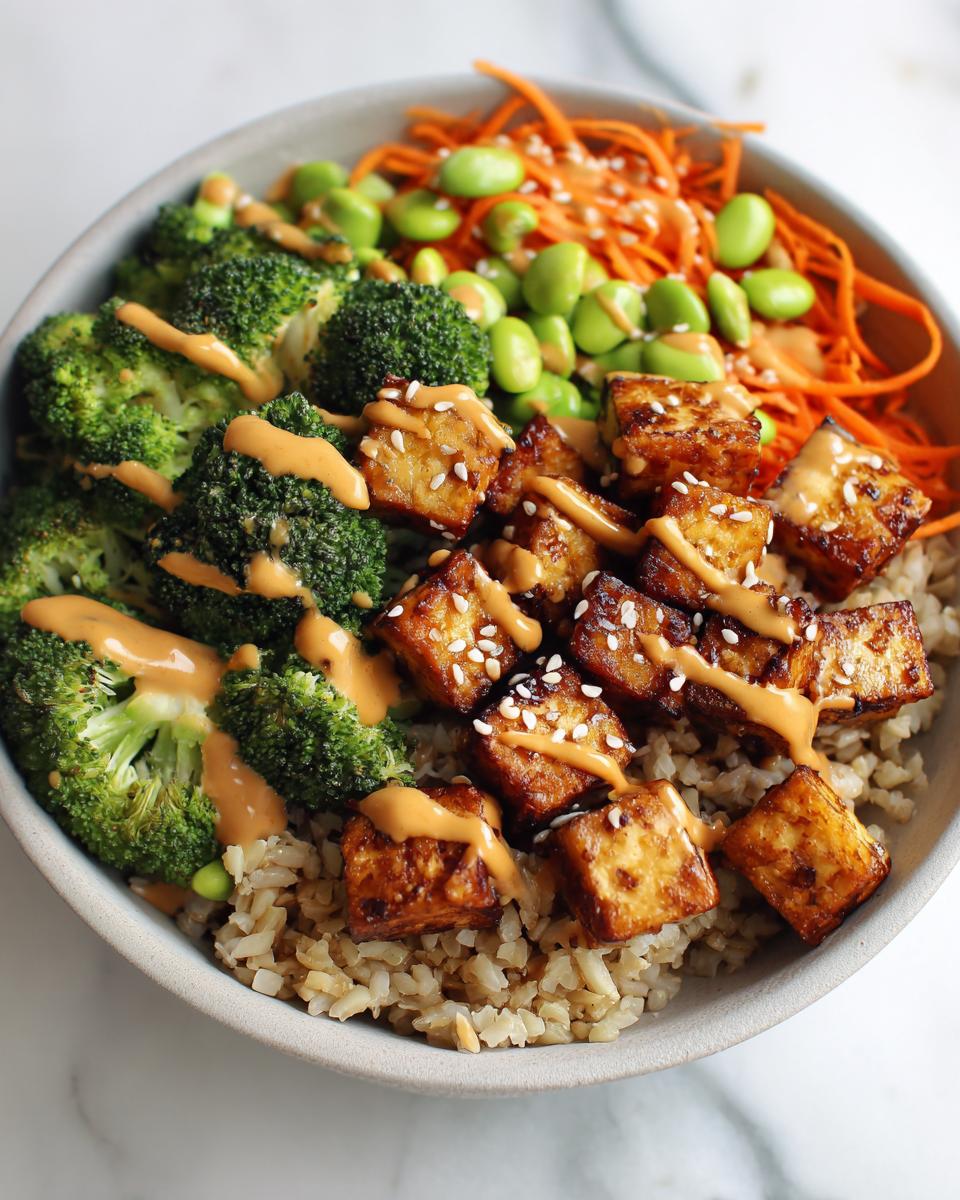 A close-up of a Crispy Tofu Buddha Bowl featuring brown rice, steamed broccoli, shredded carrots, edamame, and crispy tofu cubes drizzled with sauce.