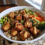 A close-up of a Crispy Tofu Buddha Bowl featuring brown rice, crispy tofu cubes, edamame, broccoli, carrots, and peanut sauce.