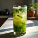 A tall glass filled with a vibrant green Cucumber Ginger Mint Mocktail, ice, and fresh mint leaves, sparkling with bubbles.