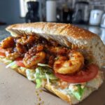 A close-up of The Best Easy Blackened Shrimp Po Boy sandwich overflowing with blackened shrimp, lettuce, and tomato on a crusty roll.