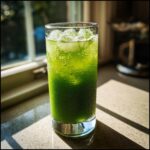 A tall, condensation-covered glass filled with a vibrant Green Goddess Mocktail and ice cubes, sitting in sunlight.