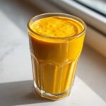 A tall glass filled with a thick, bright orange Healthy Pineapple Strawberry Turmeric Smoothie, glistening with condensation.