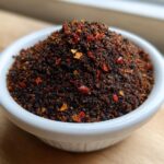A close-up of a small white bowl overflowing with dark, richly spiced Homemade Blackened Seasoning, featuring visible red pepper flakes.