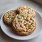 Three Loaded Lucky Charms Cornflakes Sugar Cookies topped with colorful marshmallows and cereal pieces on a white plate.