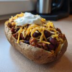 A baked potato split open and filled with spicy beans, ground meat, shredded cheese, and a dollop of sour cream, making for perfect Loaded Potatoes With Spicy Beans.