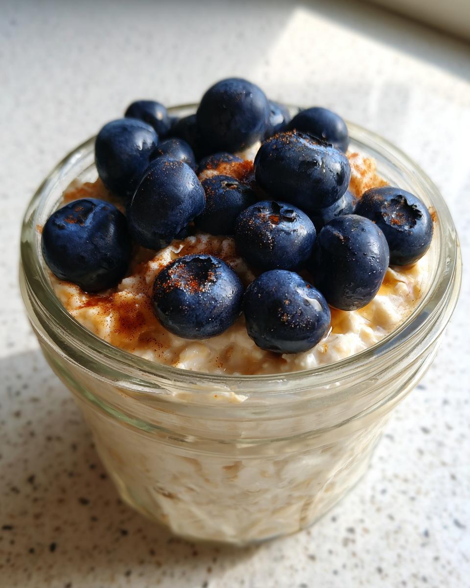 Close-up of Maple Cinnamon Overnight Oats in a glass jar, topped generously with fresh blueberries and a sprinkle of cinnamon.