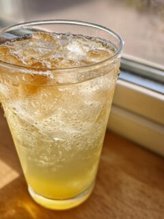 A tall, sweating glass filled with Natural Homemade Lemon Ginger Ale and ice cubes, sitting on a sunlit wooden surface.