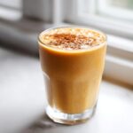 A tall glass filled with a thick, orange Peach Cobbler Smoothie, topped with a sprinkle of cinnamon.