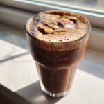 A tall glass filled with a dark brown, frothy Peanut Butter Mocha Smoothie, sitting by a sunlit window.