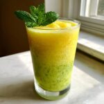 A tall glass filled with a vibrant, layered Pineapple Mint Smoothie, garnished with fresh mint leaves.