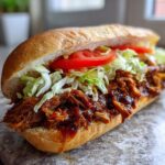 Close-up of a long roll filled with saucy BBQ Jerk Pulled Pork Sandwiches Plant Based, lettuce, and tomato.