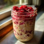A glass jar filled with layered Raspberry Coconut Overnight Oats, topped with fresh raspberries.