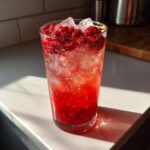 A tall glass filled with a vibrant red Raspberry Lemon Mocktail, ice cubes, and fresh raspberries, sitting on a sunlit countertop.