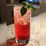 A tall glass filled with a vibrant red Strawberry Basil Pineapple Mocktail over crushed ice, garnished with fresh basil.