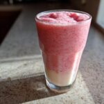 A tall glass filled with a thick, pink Strawberry Coconut Milkshake, showing a layer of white coconut milk at the bottom.