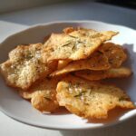 A pile of golden, crispy Sweet Spicy Herb Crackers topped with rosemary and sea salt on a white dish.