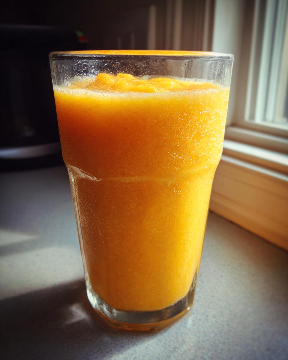 A tall glass filled with a thick, bright orange Tropical Mango Smoothie sitting on a countertop.
