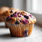 A close-up, appetizing shot of a freshly baked Vegan Blueberry Muffin with a golden top studded with purple berries.