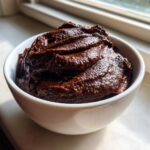 A close-up of rich, dark Vegan Chocolate Avocado Pudding scooped high in a small white bowl near a window.