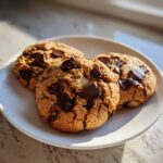Three freshly baked Vegan Chocolate Chip Cookies with melted chocolate chunks resting on a white plate.