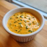 Close-up of bright orange Vegan Jalapeno Queso Dip, flecked with green herbs, served in a white ramekin.