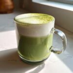 A vibrant green Vegan Matcha Latte with thick white foam, dusted with matcha powder, in a clear glass mug.