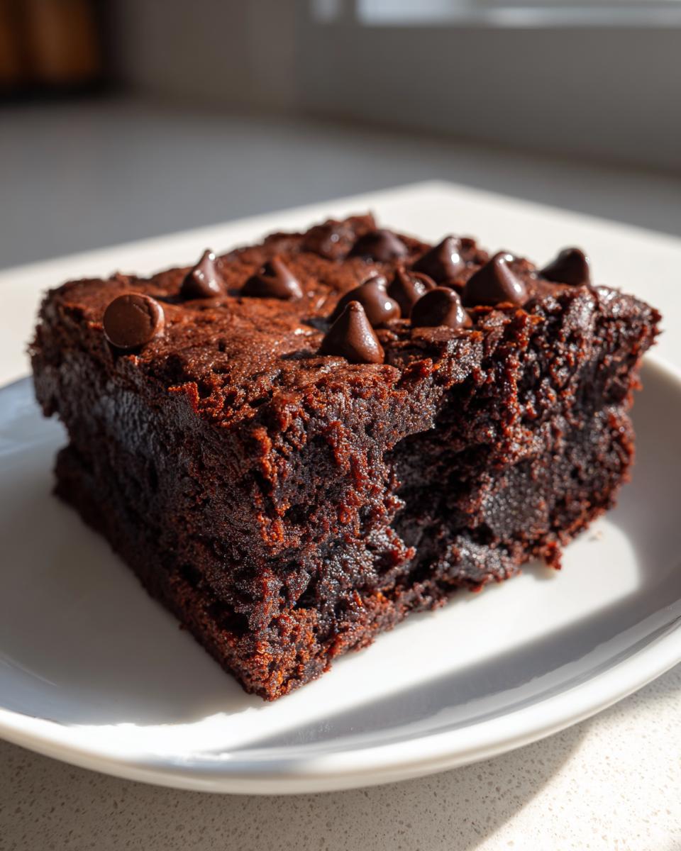 Close-up of a rich, dark Vegan Sweet Potato Brownies square topped with chocolate chips on a white plate.