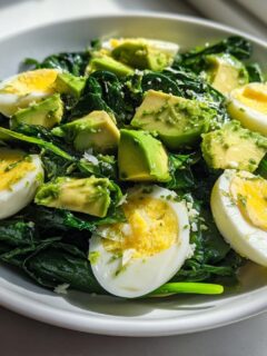 A bright, close-up photo of an Avocado Egg And Spinach Salad served in a white bowl, featuring halved hard-boiled eggs and chunks of fresh avocado.