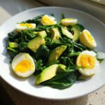 A vibrant Avocado Egg And Spinach Salad featuring sliced avocado and halved hard-boiled eggs on a bed of fresh spinach.