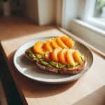 Two slices of Avocado Peach Toast topped with bright orange peach slices and pepper, sitting on a plate.