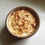 A close-up overhead shot of baked French Onion Dip in a small brown ramekin, showing a creamy texture with caramelized onions on top.