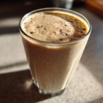 Close-up of a thick, frothy Banana Chip Smoothie in a clear glass, showing small dark flecks.